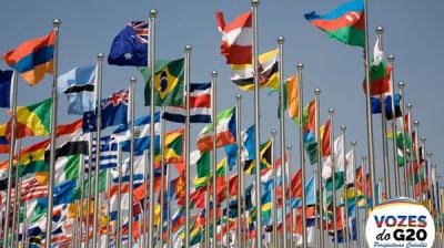 Numerous international flags on poles, waving against a clear sky. A logo in the corner reads "Vozes do G20 Perspectivas Cidadãs.