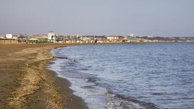 A sandy beach with gentle waves, bordered by a town with buildings and colorful beach huts in the background under a clear sky.