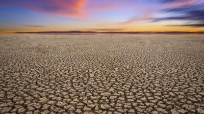 Cracked dry earth stretches toward a colorful sunset sky with pink and orange clouds on the horizon.