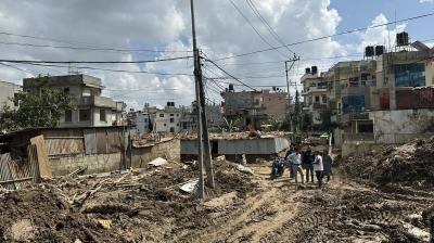 A group of people walk through a muddy, debris-filled street in a residential area, with damaged buildings and clouds in the sky.