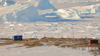 Two small structures on a barren landscape with snow and large ice formations in the background.