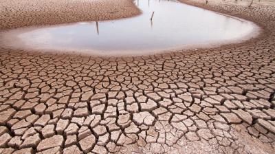 Cracked, dry earth surrounds a small pool of water in an arid landscape under a cloudy sky.