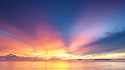 A vibrant sunset over calm waters, with a sky transitioning from orange and yellow near the horizon to deep blue above, and silhouetted islands on the right.