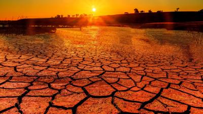 A cracked, dry earth landscape under a vibrant orange sunset, with silhouettes of distant trees along the horizon.