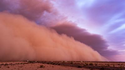 A large dust storm moves across a desert landscape beneath a colorful sky.