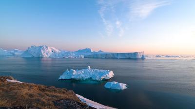 Icebergs float in a calm sea under a clear sky at sunrise, with a rocky shoreline in the foreground.