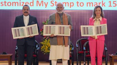 Three individuals stand holding stamp sheets at a ceremony. A sign in the background mentions "India Meteorological Department" and "Customized My Stamp.