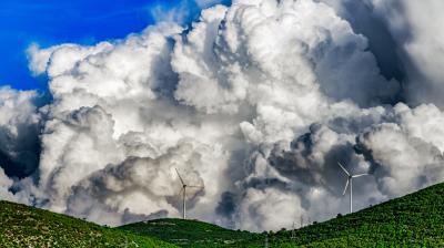 Large, dramatic clouds rise above green hills with wind turbines.
