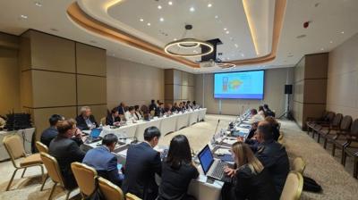 Conference room with a large group of people seated around a U-shaped table. A presentation is displayed on a screen at the front of the room.