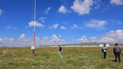 Two people stand beside a tall, slender weather station tower on a grassy field under a blue sky with white clouds.