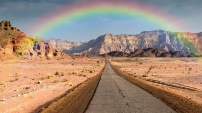 A straight road stretches through a rocky desert landscape with a rainbow arching above distant cliffs under a cloudy sky.