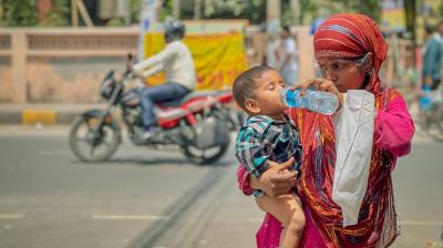 A woman in a colorful outfit gives water from a plastic bottle to a young child on a street with a motorbike passing by.