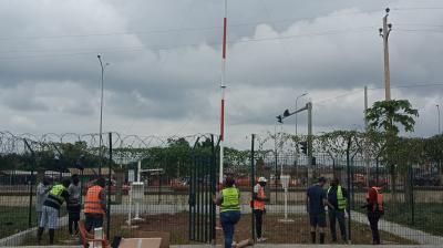 Workers in safety vests install a tall, red and white pole within a fenced area on a cloudy day. Nearby, boxes and wooden planks are on the ground.