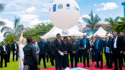 A group of people, including officials in suits, stand on a red carpet outdoors beneath a large white balloon with logos, surrounded by greenery and tents.