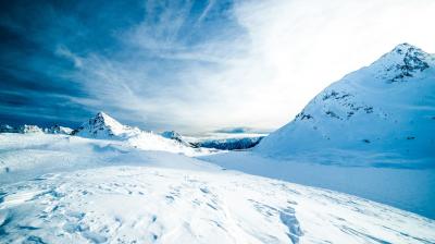 Snow-covered mountain landscape under a blue sky with scattered clouds, featuring two prominent peaks and soft shadows on the snow.