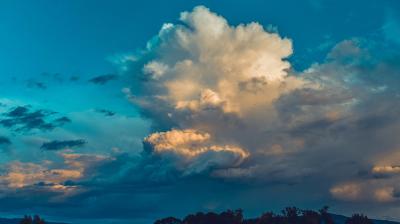 A large, billowing cloud in a blue sky over a landscape with dark treetops at the bottom.
