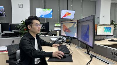 A person sits at a desk in an office with multiple monitors displaying weather maps and data.