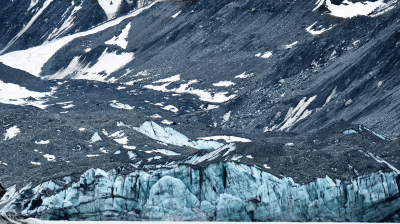 A glacier with blue ice formations is surrounded by rocky, snow-dusted mountains, and a body of water is visible in the foreground.