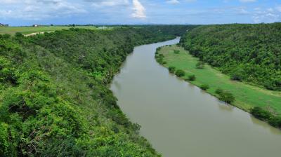 Aerial view of a wide river flowing through a lush, green valley, surrounded by dense forest and grassy slopes under a blue, cloud-dotted sky.
