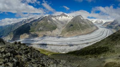 Panoramic view of a glacier winding between rugged mountains under a blue sky with scattered clouds. Rocky foreground leads to the expansive ice flow, surrounded by green slopes and rocky peaks.