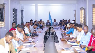 A large group of people in a meeting room, sitting at a long table facing forward. A flag is positioned at the end of the table. Many have laptops open.
