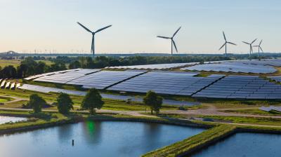 A landscape featuring a solar farm and multiple wind turbines on a grassy area, with ponds in the foreground and a clear sky in the background.