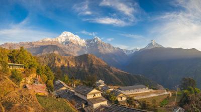 A Himalayan village with traditional houses sits in the foreground, surrounded by green hills. Snow-capped mountains rise under a clear blue sky in the background.