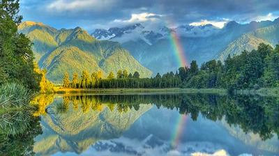 A calm lake reflects surrounding mountains, trees, clouds, and a rainbow in the sky.