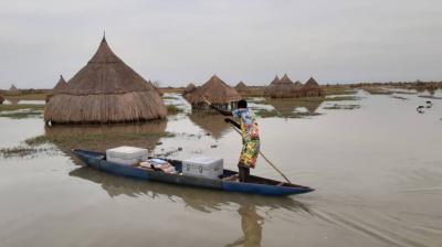 A person paddles a small canoe on a floodplain, passing traditional thatched huts partially submerged in water.