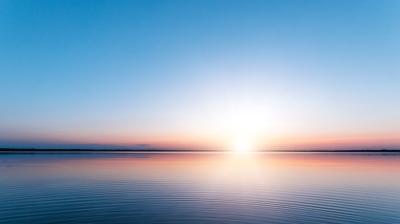 A calm body of water reflects the pastel colors of a sunrise or sunset, with a horizon line separating the sky from the water.