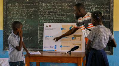 Teacher explaining a safety poster to two students in front of a chalkboard in a classroom. The chalkboard is filled with text, and a microphone is visible on the table.