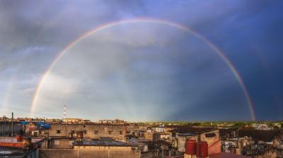 A panoramic view of a cityscape under a dark, cloudy sky, with a vibrant double rainbow arching above the buildings.