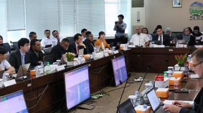 A large group of people in a conference room sit around a U-shaped table with microphones, laptops, and papers. A man stands at the back with a camera. Screens display information.