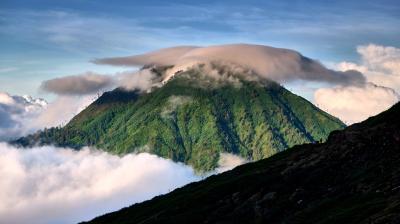 A green mountain peak is partially covered by a cap of clouds, with additional clouds surrounding its base under a clear blue sky.