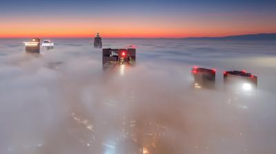 Tall city buildings rise above dense fog at sunrise, with the sky displaying a gradient from deep blue to orange on the horizon.