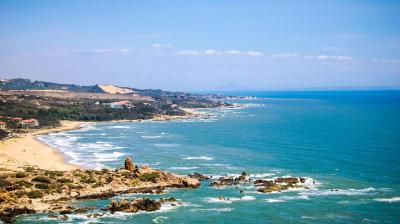 Coastal landscape with sandy beaches, rocky shoreline, and gentle waves under a blue sky, with hills and scattered buildings in the background.