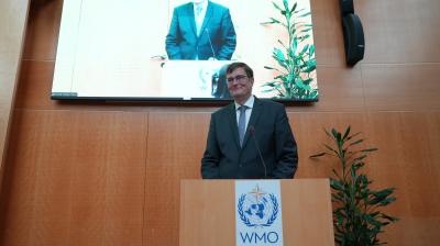 A man in a suit stands at a podium with the WMO logo, speaking in front of a screen displaying his image in a wood-paneled room with plants.