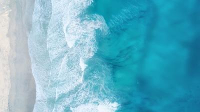 Aerial view of waves crashing onto a sandy beach, with clear blue water and white surf along the shoreline.