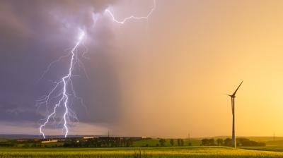 A lightning bolt strikes in the distance over fields at sunset, with a single wind turbine standing in the foreground.