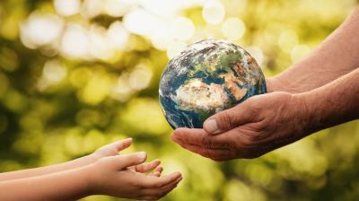 Two adult hands hold a small globe while two child hands reach out towards it, set against a blurred, green outdoor background.