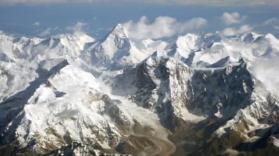 Aerial view of snow-covered mountain peaks with a glacier winding through the valley below under a partly cloudy sky.