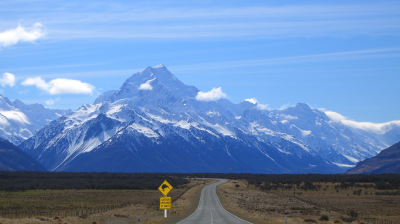 A straight road leads toward snow-covered mountains under a blue sky, with a yellow road sign on the left side of the road in the foreground.