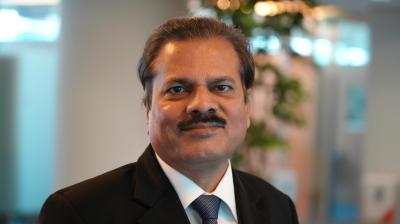 A middle-aged man in a suit and tie with a small Indian flag pin, standing indoors with blurred office background.