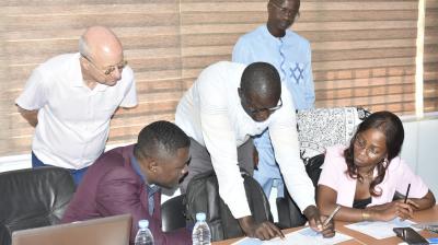 Five people in an office setting discuss documents spread out on a table, with laptops, water bottles, and papers visible.