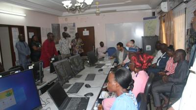 A group of people sits and stands around computer workstations in a classroom or office setting during a meeting or training session.