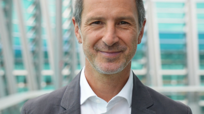 A middle-aged man with short dark hair and a beard smiles at the camera, wearing a white shirt and gray blazer, standing in front of a modern glass building.
