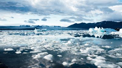Large chunks and sheets of ice float on a calm, reflective body of water, with mountains and a cloudy sky in the background.