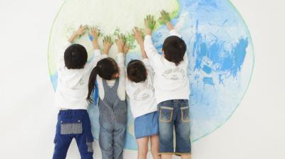 Four children stand facing a wall, pressing their painted hands onto a large drawing of the Earth.