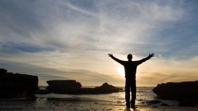 A person stands on a rocky beach with arms outstretched, facing the ocean at sunset under a partly cloudy sky.