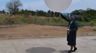 A person in a coat and headscarf stands on a paved area holding a large white weather balloon outdoors under a blue sky.